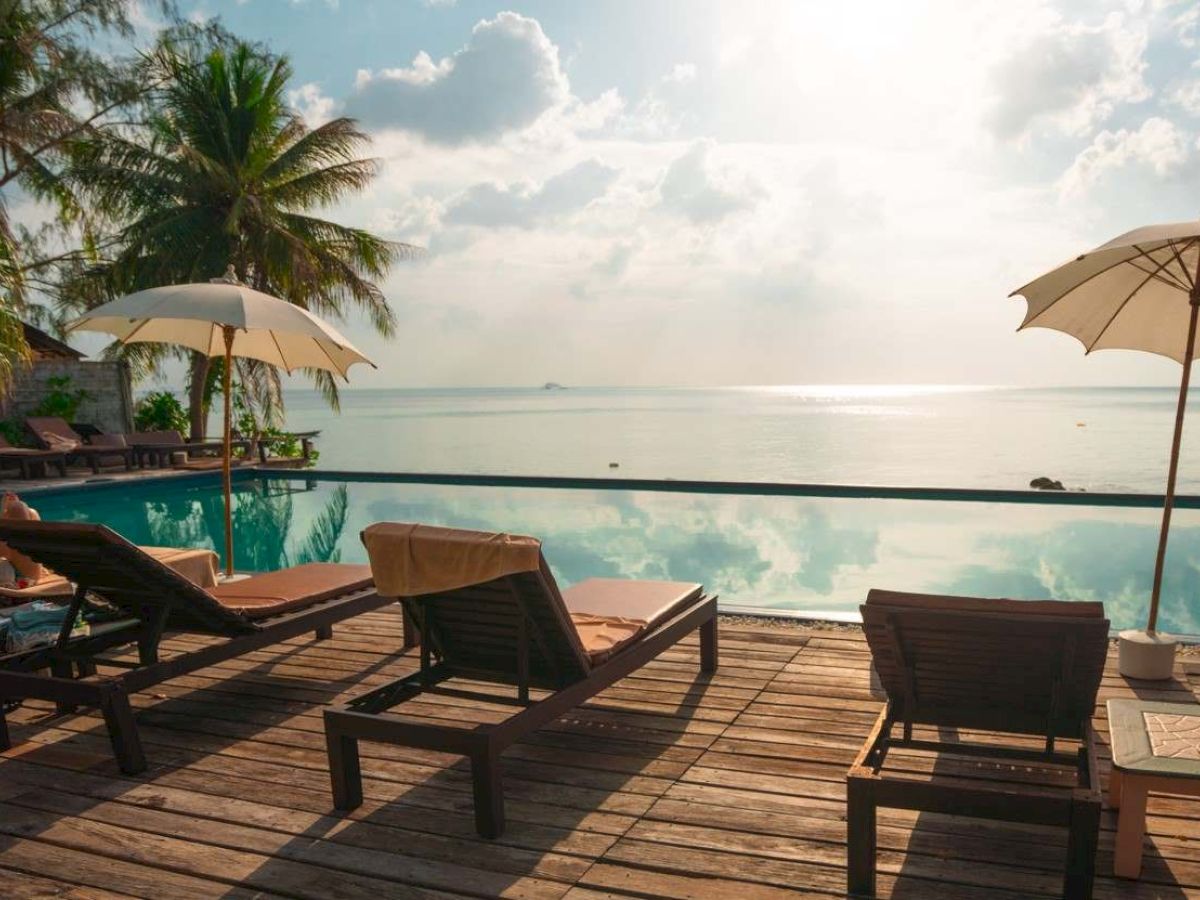 Lounge chairs and umbrellas by a calm poolside, overlooking a serene ocean view, surrounded by palm trees under a partly cloudy sky.