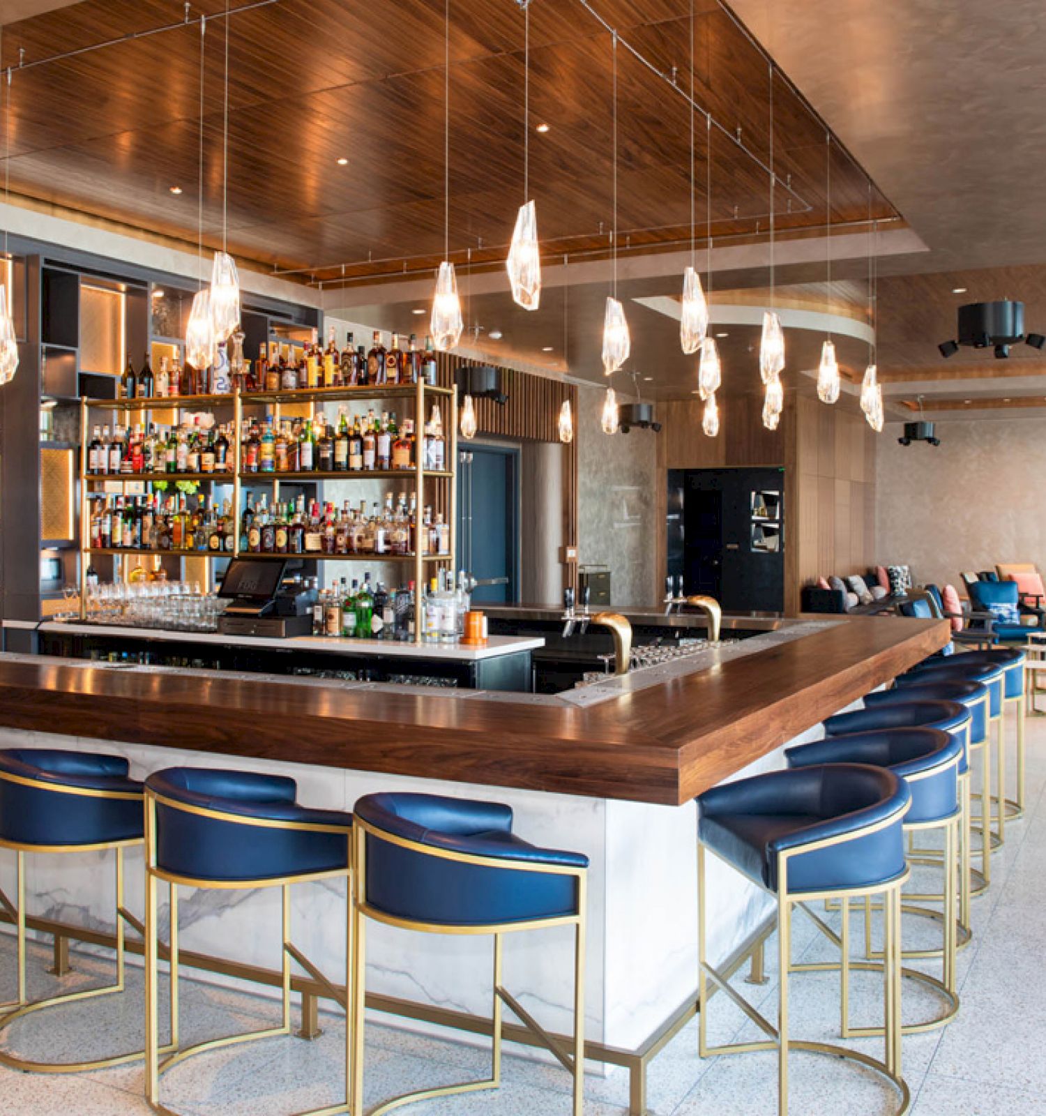 The image shows a modern bar and lounge area with a counter, pendant lights, and cushioned bar stools. Shelves display bottles behind the bar.
