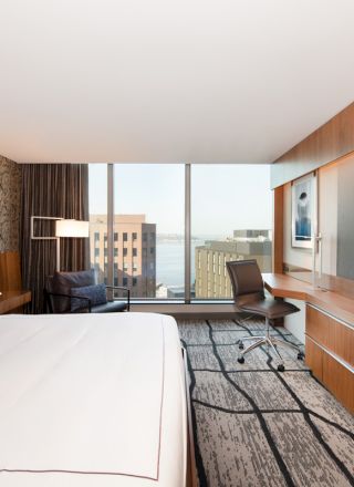 The image shows a modern hotel room with a large bed, a desk, a TV, and a window with a city view in the background.