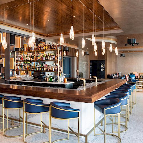 The image shows a modern bar with a wooden counter, blue barstools, pendant lights, and a well-stocked back shelf in an elegant lounge setting.