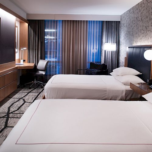 The image shows a modern hotel room with two beds, a TV, a work desk, and stylish lighting and decor.
