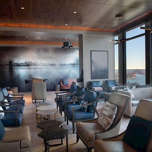 A modern lounge with plush seating, large windows, and a view of water and a Ferris wheel, under warm lighting and wall art.