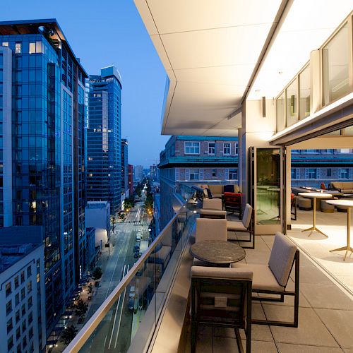 A modern rooftop terrace with tables and chairs overlooks a cityscape with tall buildings at dusk, offering a serene urban view.
