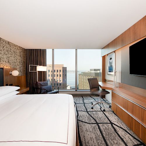 A modern hotel room features a large bed, sleek desk, flat-screen TV, and a window view of buildings and water.