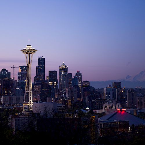 A cityscape of Seattle at dusk with the Space Needle, skyline, and Mount Rainier in the background under a twilight sky.