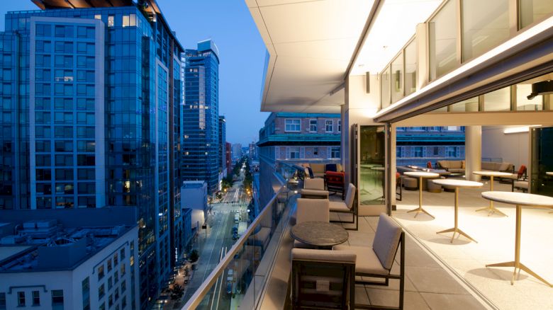A modern balcony overlooking a cityscape at dusk, with outdoor seating and bright interior lights visible through large windows.