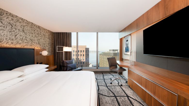 A modern hotel room with a large bed, flat-screen TV, desk, chair, and a window with a city view.