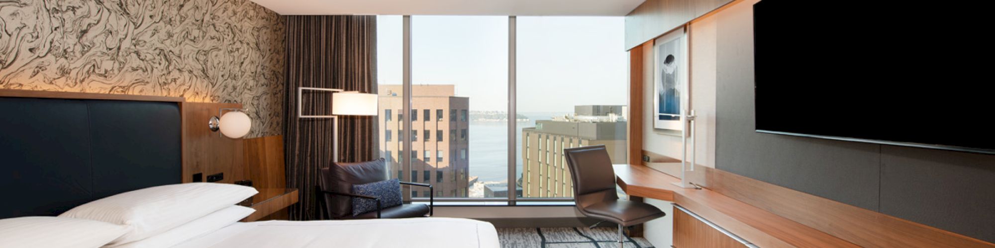 A modern hotel room with a large bed, desk, chair, TV, and a window showing a city view in the daylight.